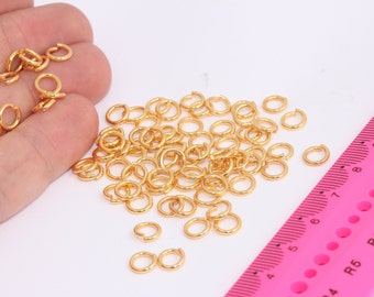 May include: A large pile of gold jump rings, a type of jewelry finding, are scattered on a white surface. A pink ruler with black markings is in the lower right corner of the image.