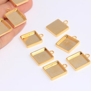 May include: Gold-coloured square pendant trays with a small loop for attaching to a chain or other jewellery component. The trays have a recessed area for setting a design or image. Several are visible.