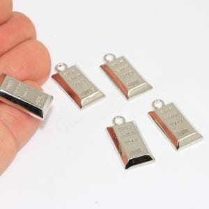 Puede incluir: Encantos rectangulares plateados con forma de lingotes de oro. Cada dije tiene un pequeño lazo en la parte superior para sujetarlo a una joya. Los dijes están grabados con texto y se muestran sobre un fondo blanco.