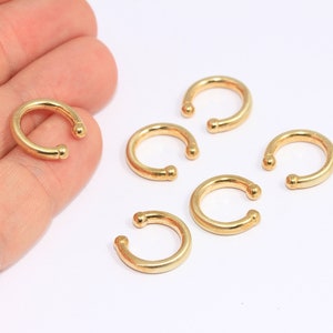 Könnte beinhalten: Mehrere goldfarbene Ohrklemmen werden auf einer weißen Oberfläche gezeigt. Die Ohrklemmen sind kreisförmig mit einer kleinen Kugel an jedem Ende der Öffnung. Eine Manschette wird in einer Hand gehalten. Dies sind Schmuckaccessoires.