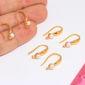 Könnte beinhalten: Goldfarbene Ohrringhaken mit einem kleinen, weißen Perlenakzent. Die Haken sind gebogen und haben eine Schlaufe zum Anbringen von Anhängern. Mehrere Haken sind sichtbar.