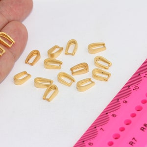 Puede incluir: Primer plano de 17 baúles de colgante de metal dorado, que son pequeños bucles utilizados para sujetar colgantes a collares o pulseras. Los baúles están esparcidos sobre una superficie blanca junto a una regla rosa.