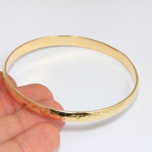 Könnte beinhalten: Ein goldfarbenes Armreif-Armband mit gehammerter Textur. Das Armband ist rund und scheint aus Metall zu sein. Das Armreif wird in der Hand einer Person vor einem weißen Hintergrund gehalten. Das Armband hat einen Durchmesser von etwa 6,35 cm.