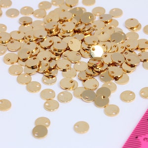 Puede incluir: Un montón grande de dijes redondos de metal dorado con un pequeño agujero en el centro. Los dijes están esparcidos sobre una superficie blanca junto a una regla rosa.