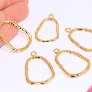May include: Five gold-toned metal pendant charms with an irregular oval shape and a small loop at the top. The charms are arranged on a white surface.
