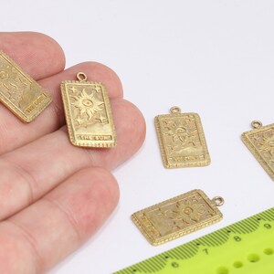 Könnte beinhalten: Fünf goldfarbene Metall-Anhänger in Form von Tarotkarten, jede mit dem Design der Karte "Die Sonne". Die Anhänger liegen auf einer weißen Oberfläche mit einem grünen Lineal im Hintergrund.