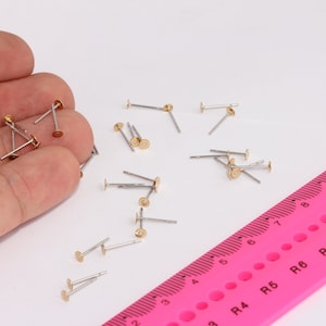 Peut inclure: Gros plan sur une main tenant un tas de petites tiges d'oreille en métal de couleur dorée. Les tiges sont éparpillées sur une surface blanche à côté d'une règle rose avec des marques noires.