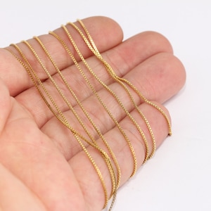 Op de afbeelding: Een close-up van een gouden ketting. De ketting is gemaakt van kleine, met elkaar verbonden vierkanten. De ketting ligt op een hand.