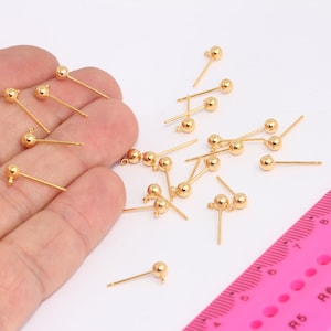 May include: A close-up of a pile of gold-colored earring findings with a round ball at the top. The findings are scattered on a white surface next to a pink ruler.