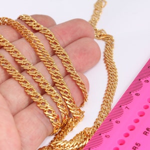 Peut inclure: Un collier en chaîne dorée avec un design de maillons curb. La chaîne est posée sur une main et à côté d'une règle rose avec des marques noires.