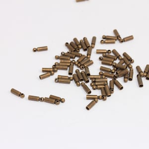 Puede incluir: Una gran cantidad de pequeños tapones metálicos de color dorado con una bola en un extremo. Los tapones están dispersos sobre una superficie blanca.