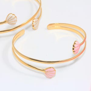 Pode incluir: Pulseiras de punho douradas com detalhes florais. As pulseiras apresentam um design de faixa dupla e são adornadas com detalhes em forma de flor esmaltada em branco e rosa. Uma régua rosa está no fundo.