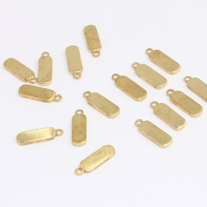 Può includere: Un gruppo di 14 ciondoli rettangolari in metallo dorato con un anello in cima. I ciondoli sono sparsi su una superficie bianca.