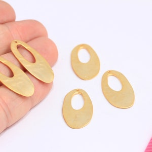 Puede incluir: Cuatro colgantes de pendientes dorados en forma ovalada con un diseño recortado. Dos se sostienen en una mano y dos están sobre una superficie blanca. Una regla rosa está en el fondo.