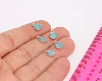 May include: A collection of small, round silver charms with a turquoise stone design. Each charm has a small loop at the top. A pink ruler is in the background.
