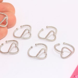 Puede incluir: Seis anillos de plata en forma de corazón con un diseño de cuerda trenzada. Los anillos están dispuestos en semicírculo sobre una superficie blanca.