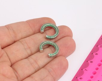 May include: A pair of silver hoop earrings with green cubic zirconia stones. The earrings are shown on a hand.