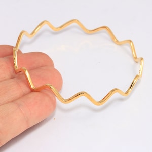 Op de afbeelding: Een golvende, open armband van goudkleur. De armband is gemaakt van een dun, flexibel metaal en heeft een eenvoudig, modern ontwerp.