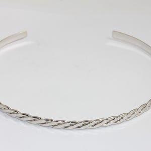 May include: A silver-colored necklace with a braided design on the front. The necklace is open at the back and has a smooth, polished finish. The jewelry is displayed on a white surface.