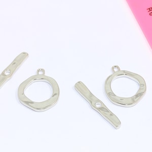 Puede incluir: Cierres de palanca plateados, ideales para la fabricación de joyas, se muestran sobre un fondo blanco. Los cierres incluyen una barra con un agujero y un anillo texturizado. Una regla rosa está en el fondo.