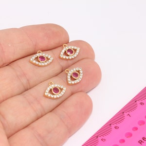 Könnte beinhalten: Vier goldfarbene böse Augen-Anhänger mit rosafarbenen Steinen und klaren Strasssteinen. Die Anhänger liegen auf einer Hand mit einem rosa Lineal im Hintergrund.