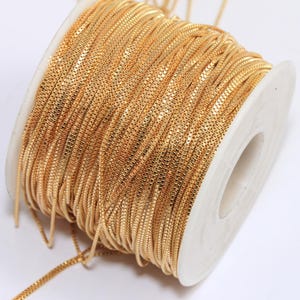 Puede incluir: Un carrete de cadena de color dorado, enrollado firmemente alrededor de un carrete de plástico blanco. La cadena es de metal fino, delicado y brillante.