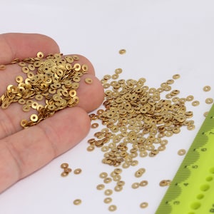 Puede incluir: Un montón grande de anillos de metal de color dorado, algunos de los cuales están dispersos sobre una superficie blanca. Una regla verde con marcas negras es visible en el fondo.