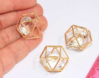 Puede incluir: Tres colgantes dorados con forma de jaula geométrica, cada uno con un cristal transparente facetado. Un colgante se sostiene en una mano, mientras que los otros dos están sobre una superficie blanca. Una regla rosa está en el fondo.