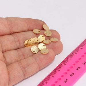 Könnte beinhalten: Nahaufnahme einer Hand, die einen Haufen kleiner, goldfarbener Metallscheiben mit zwei Löchern in jeder Scheibe hält. Die Scheiben haben einen Durchmesser von etwa 1 cm.