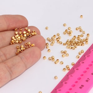 Può includere: Piccole perline dorate a forma di cubo sparse su una superficie bianca. Alcune perline sono tenute in mano. Un righello rosa con segni neri sullo sfondo, che indica le misure in centimetri.