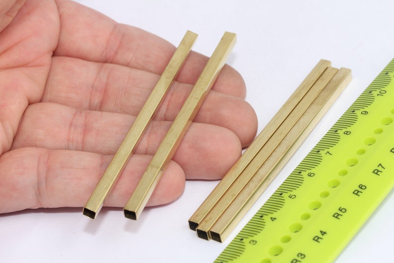 May include: Five pieces of square brass bar stock, measuring approximately 6 inches in length, are arranged on a hand and next to a ruler. The ruler is marked in inches and has a green background.