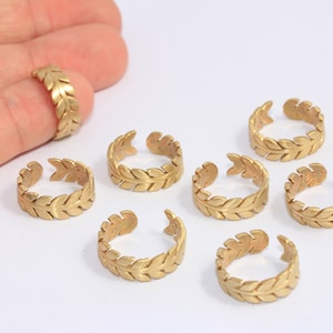 Pode incluir: Anéis ajustáveis ​​dourados com um design de folhas de louro. Os anéis são abertos e apresentam um padrão de folhas detalhado. Um anel está em um dedo e vários outros estão dispostos em uma superfície branca.