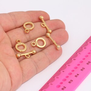 Op de afbeelding: Vijf goudkleurige metalen sluitingen voor het maken van sieraden. De sluitingen hebben de vorm van een cirkel met een staaf die door de cirkel glijdt. De sluitingen liggen op een wit oppervlak.