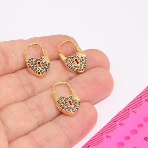 Peut inclure: Trois boucles d'oreilles en forme de cœur en or avec des pierres en oxyde de zirconium noir. Les boucles d'oreilles sont présentées sur une main avec une règle rose pour l'échelle.