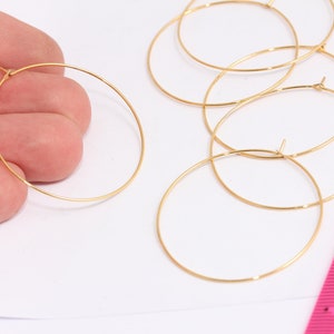 Op de afbeelding: Verschillende goudkleurige oorringen, dun en cirkelvormig, worden getoond tegen een witte achtergrond. Eén oorring wordt door een hand vastgehouden. Een roze liniaal is zichtbaar. Ideaal voor het maken van sieraden.