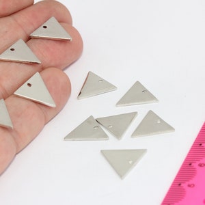 Pode incluir: Uma coleção de oito pingentes triangulares prateados com um pequeno furo no topo. Os pingentes estão dispostos sobre uma superfície branca com uma régua rosa no fundo.