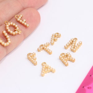 Peut inclure: Un ensemble de breloques en forme de lettres dorées avec des accents de perles blanches. Les lettres sont U, V, Y, T, M, R et A.