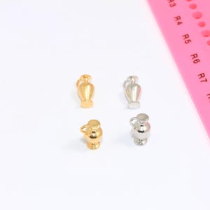 May include: Four miniature vase charms, two gold and two silver, are arranged on a white surface. A pink ruler is in the background. The charms are detailed with handles and lids, resembling small amphorae. These charms are suitable for jewellery making.
