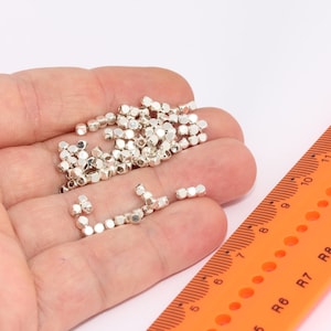 Könnte beinhalten: Nahaufnahme einer Hand, die einen Haufen kleiner, silberner, würfelförmiger Perlen hält. Im Hintergrund ist ein Lineal mit orangenen Markierungen zu sehen.