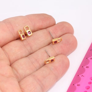 May include: A pair of gold padlock earrings with small pink gemstones. The earrings are shown on a hand with a pink ruler for scale.