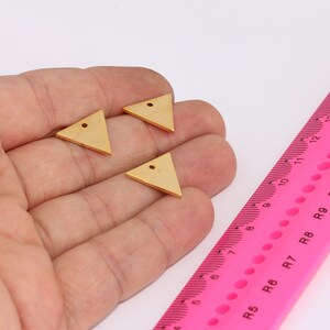 Pode incluir: Três pingentes de metal de forma triangular dourada com um furo no centro. Os pingentes medem aproximadamente 2,5 cm e são mostrados em uma mão.