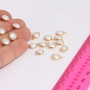 Op de afbeelding: Een verzameling witte parelbedels met goudkleurige springringen. De bedels zijn verspreid over een wit oppervlak met een roze liniaal op de achtergrond.