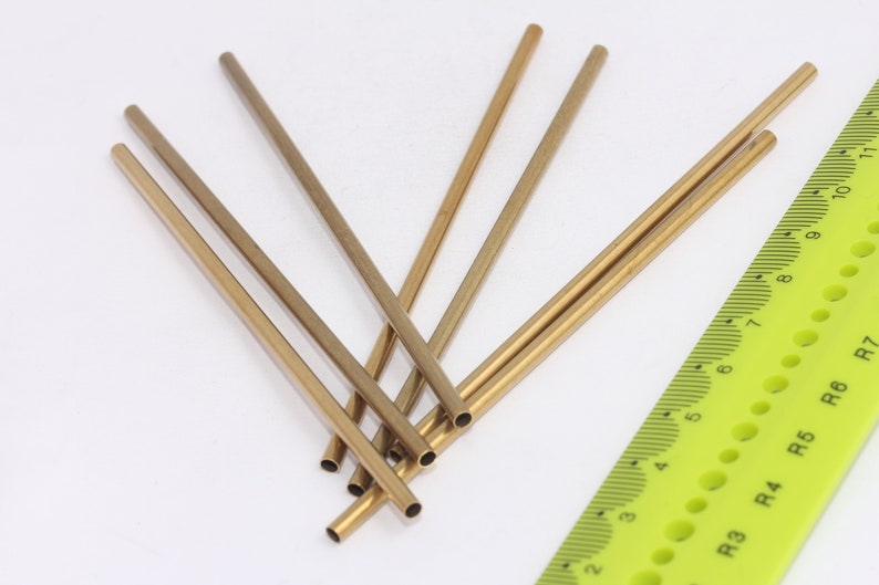 May include: Six gold-coloured metal rods of varying lengths, arranged in a starburst pattern on a white surface. A green ruler with black markings is visible in the bottom right corner of the image.