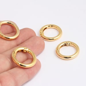 May include: Four gold-colored metal clasps, also known as trigger snaps, are shown in a hand. The clasps are round and have a spring-loaded mechanism.