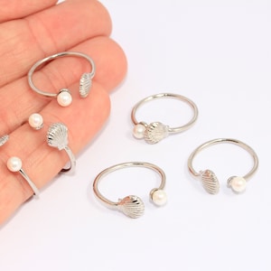 May include: Five silver rings with a pearl and a seashell design. The rings are on a white surface.