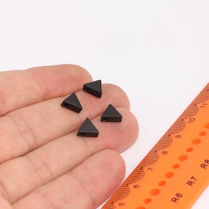 May include: Four black triangle-shaped metal beads with a hole in the centre. The beads are on a hand, next to a ruler.