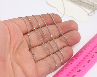 May include: Five silver chains with small, flat, square-shaped links. The chains are laid out on a hand, with a pink ruler in the background.