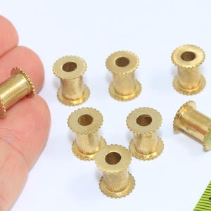Pu&ograve; includere: Una collezione di piccole perline cilindriche in ottone con un bordo scanalato e un foro al centro. Le perline sono lunghe circa 1,3 cm e sono esposte su una superficie bianca.