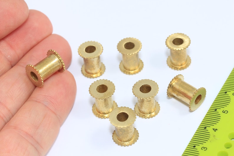 Pu&ograve; includere: Una collezione di piccole perline cilindriche in ottone con un bordo scanalato e un foro al centro. Le perline sono lunghe circa 1,3 cm e sono esposte su una superficie bianca.