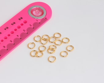 May include: A close-up of a pink ruler with a magnifying glass and gold jump rings. The ruler has markings in inches and the jump rings are scattered on a white surface.